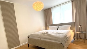 A neatly made bed with rolled towels, a bedside lamp, and a hanging light fixture in a modern room with wooden flooring and large windows.