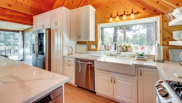 A modern kitchen with white cabinets, stainless steel appliances, and a large window providing natural light. - Wraparound Deck & Grill: Wrightwood Cabin Near PCT (Wrightwood)