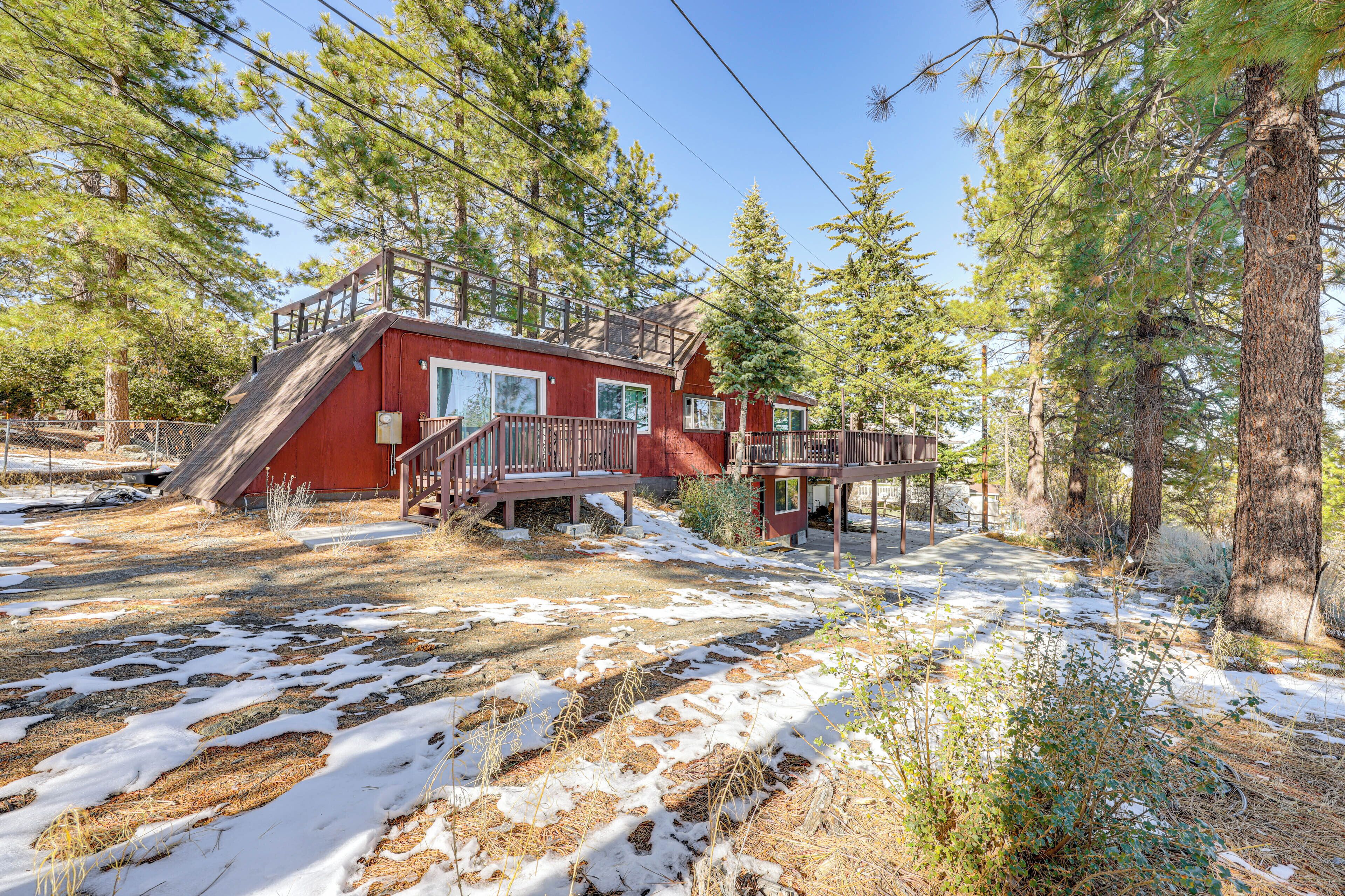 A red cabin with a wooden deck and a small porch, surrounded by trees and snow.