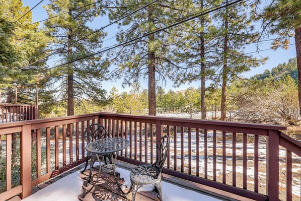 A wooden deck with metal chairs and a table, overlooking a snowy landscape with pine trees.