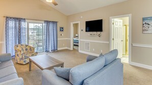 Een ruime woonkamer met een plafondventilator, een grote flatscreen-tv aan de muur en uitzicht buiten door een schuifdeur.