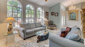 A spacious living room with a grey sofa, a glass coffee table, and a patterned armchair. There are large windows with arched tops, a ceiling fan, and a chandelier. - 10 Mi to Beach: Pet-friendly Home in Punta Gorda! (Punta Gorda)