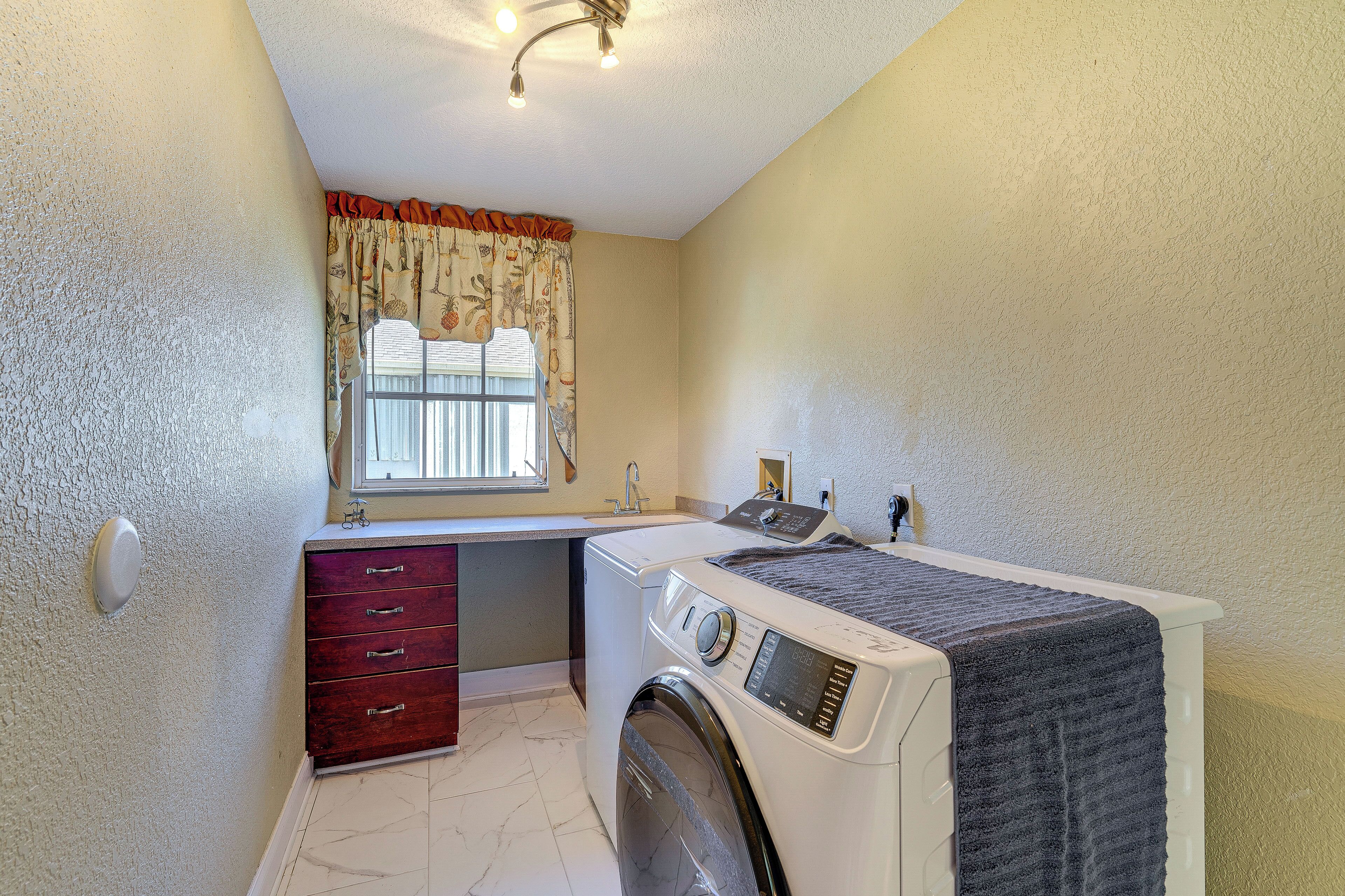 A compact laundry room with a washing machine, a dryer, a small sink, and a window with curtains.