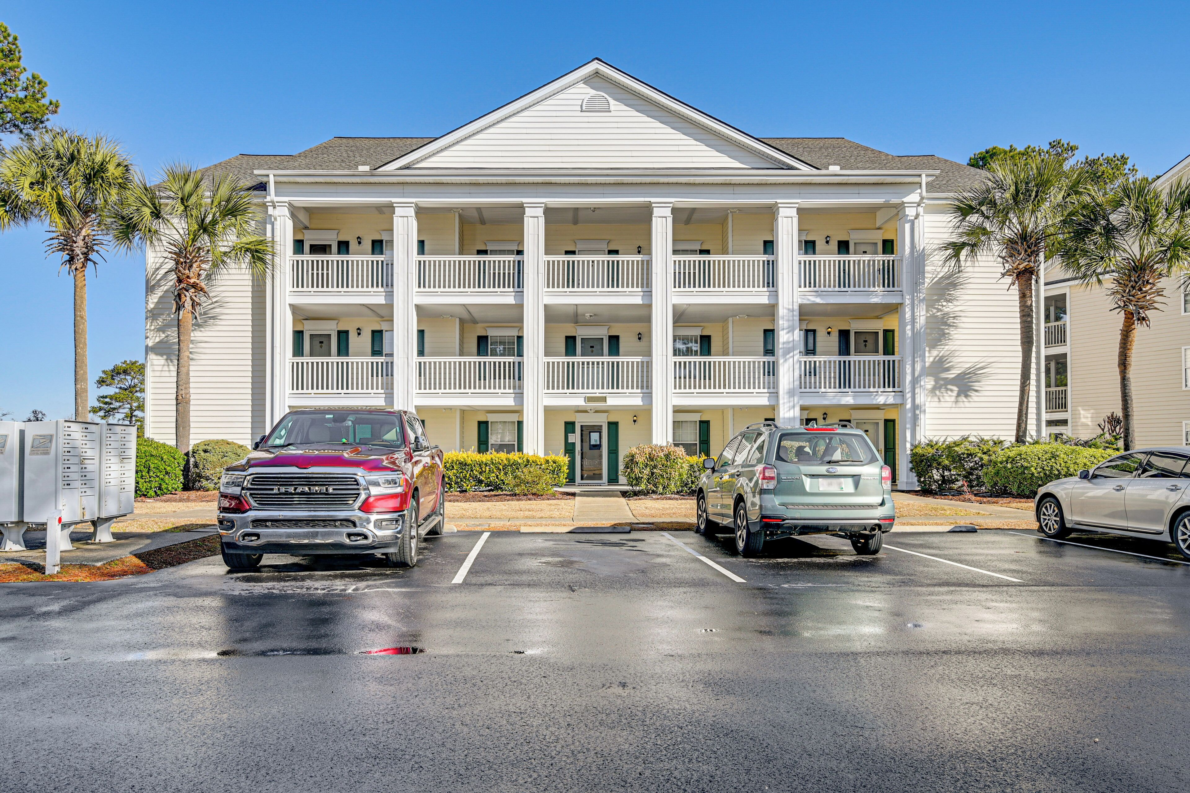 A multi-story building with a central entrance, two-story balconies, and a parking lot with three cars.
