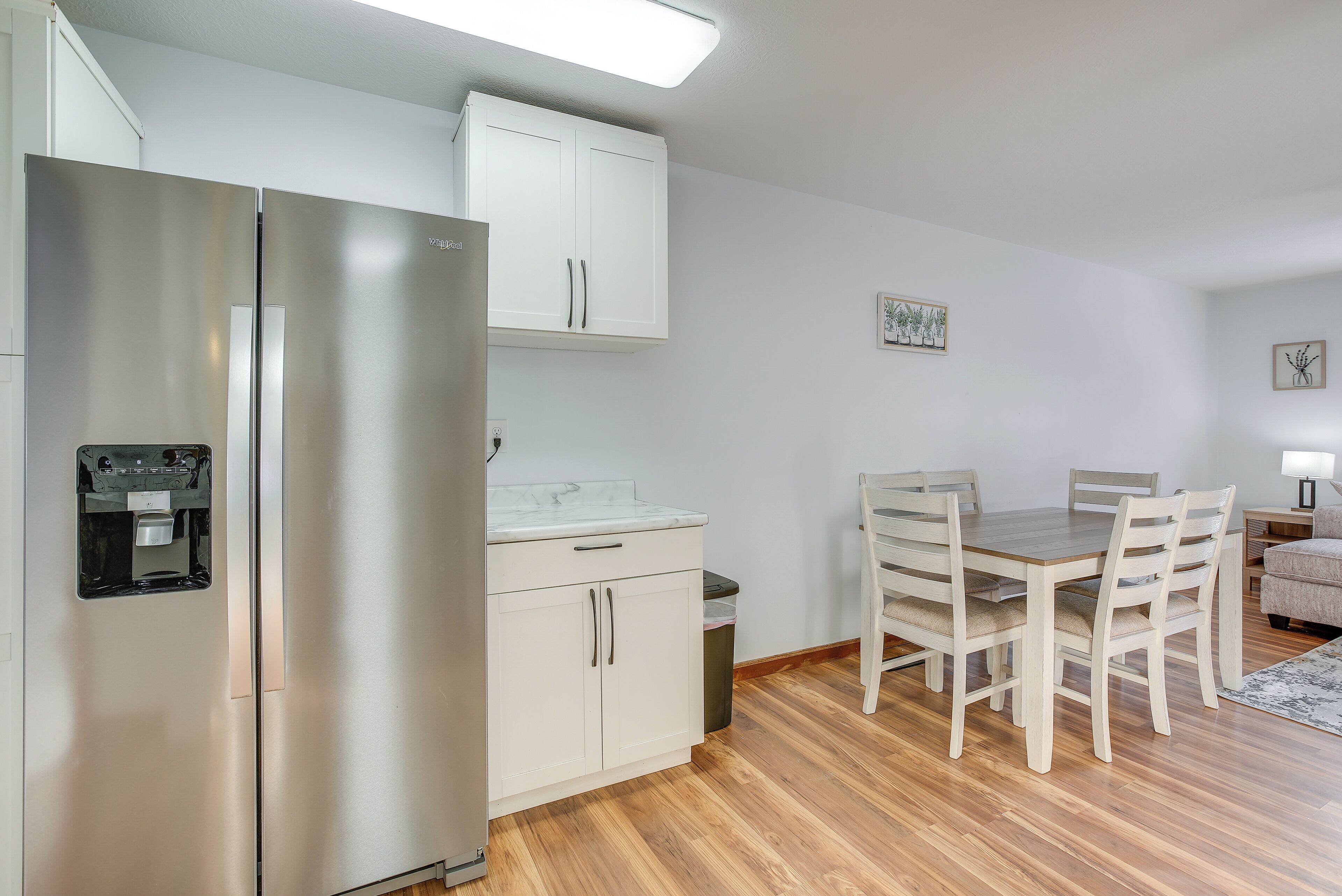 A compact kitchen with a stainless steel refrigerator, a built-in water dispenser, a white countertop, and a dining area with a wooden table and chairs.