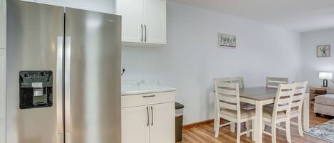 A compact kitchen with a stainless steel refrigerator, a built-in water dispenser, a white countertop, and a dining area with a wooden table and chairs.