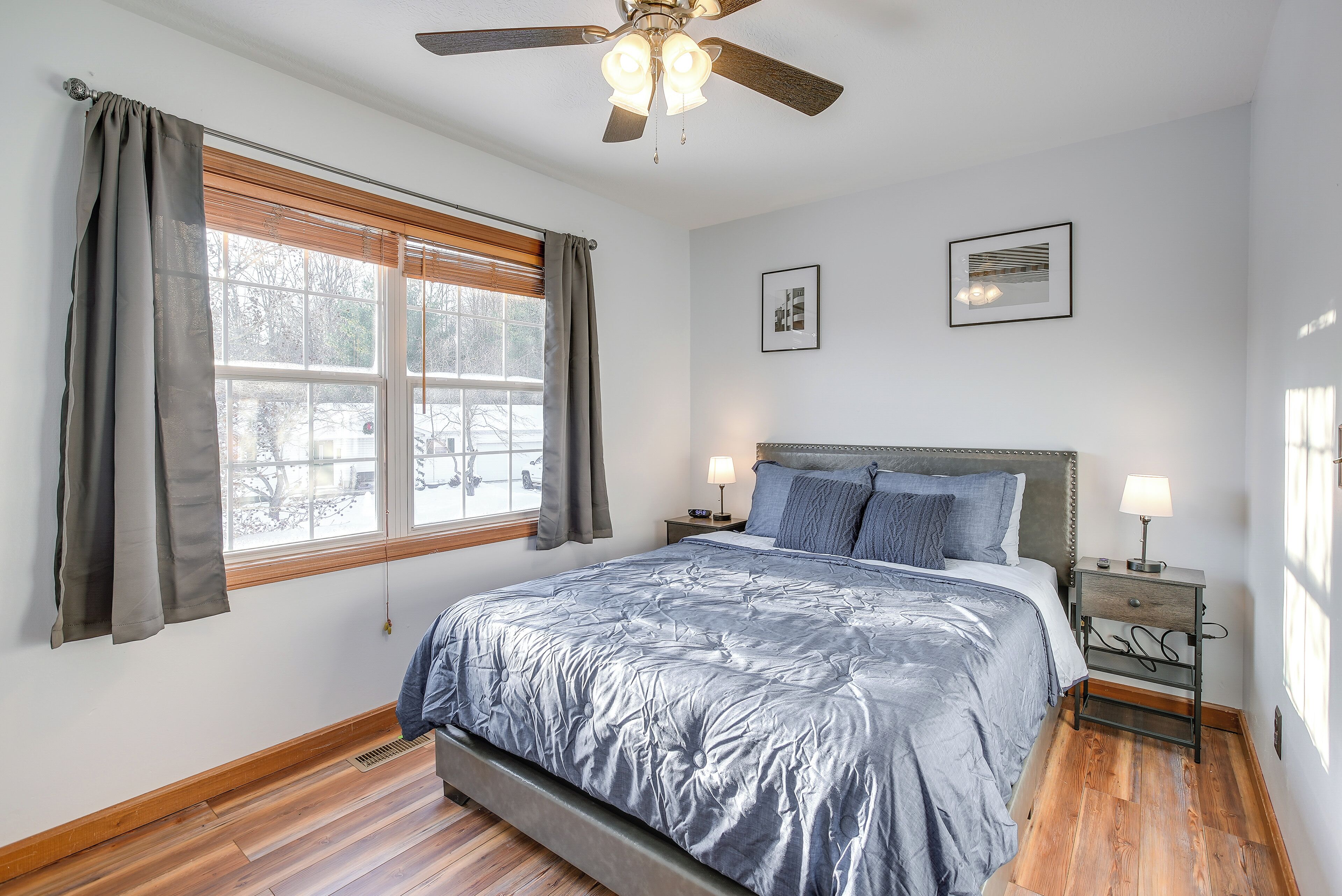 A bedroom with a large bed, a ceiling fan, a nightstand with a lamp, and a window with curtains.