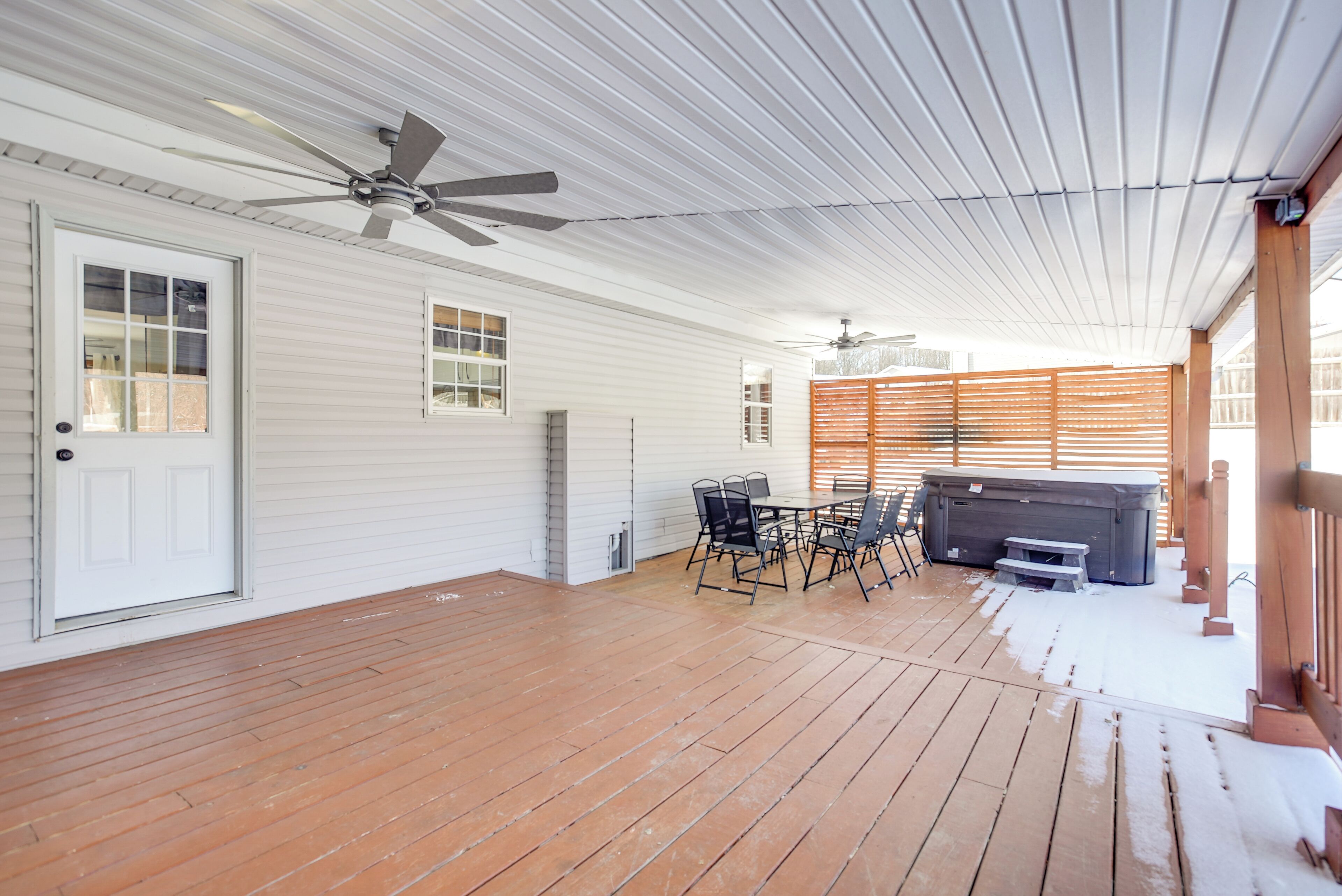 A covered patio with a wooden deck, a dining table with chairs, a hot tub, and ceiling fans.