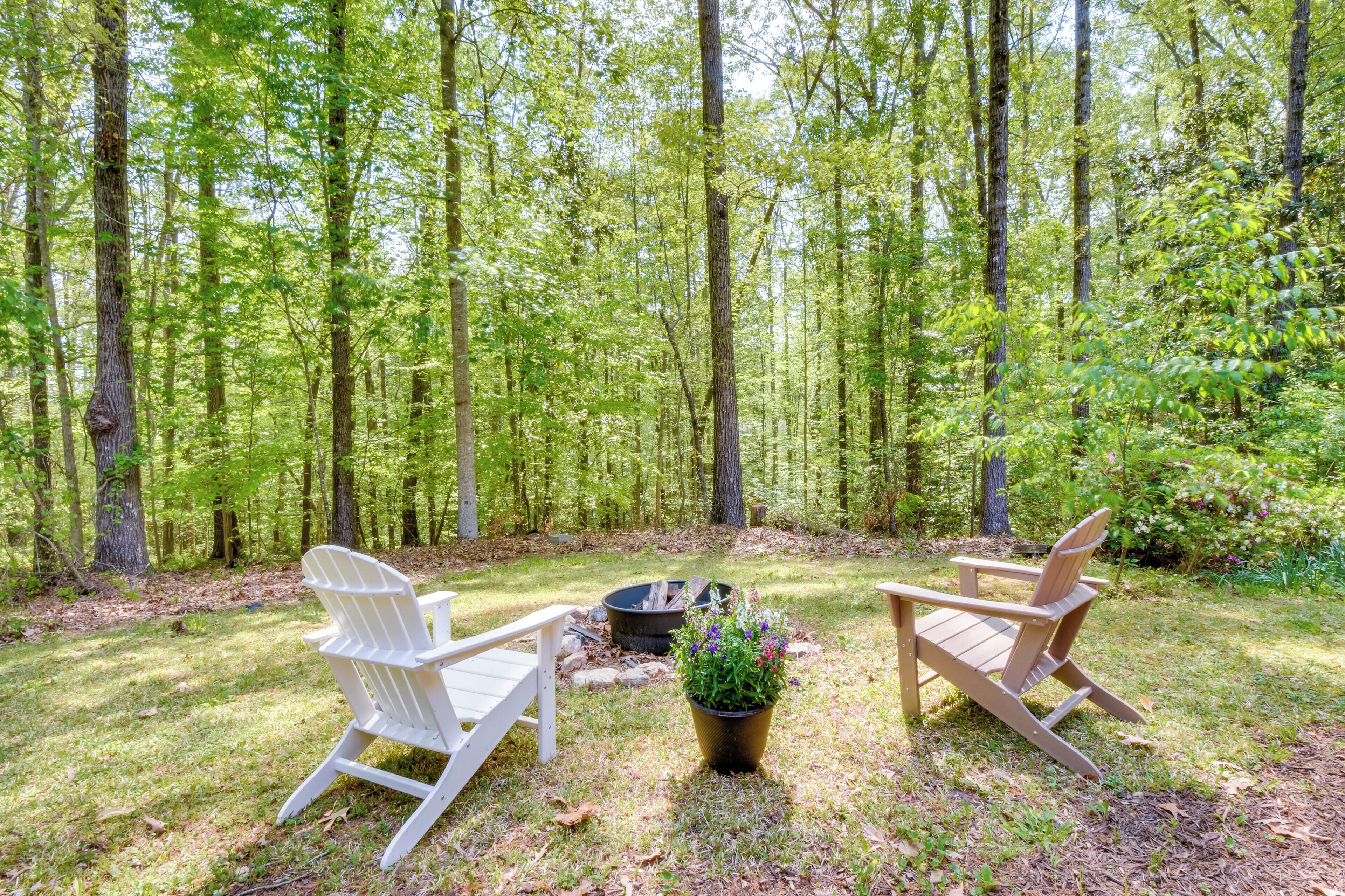 A forested area with two Adirondack chairs and a fire pit.