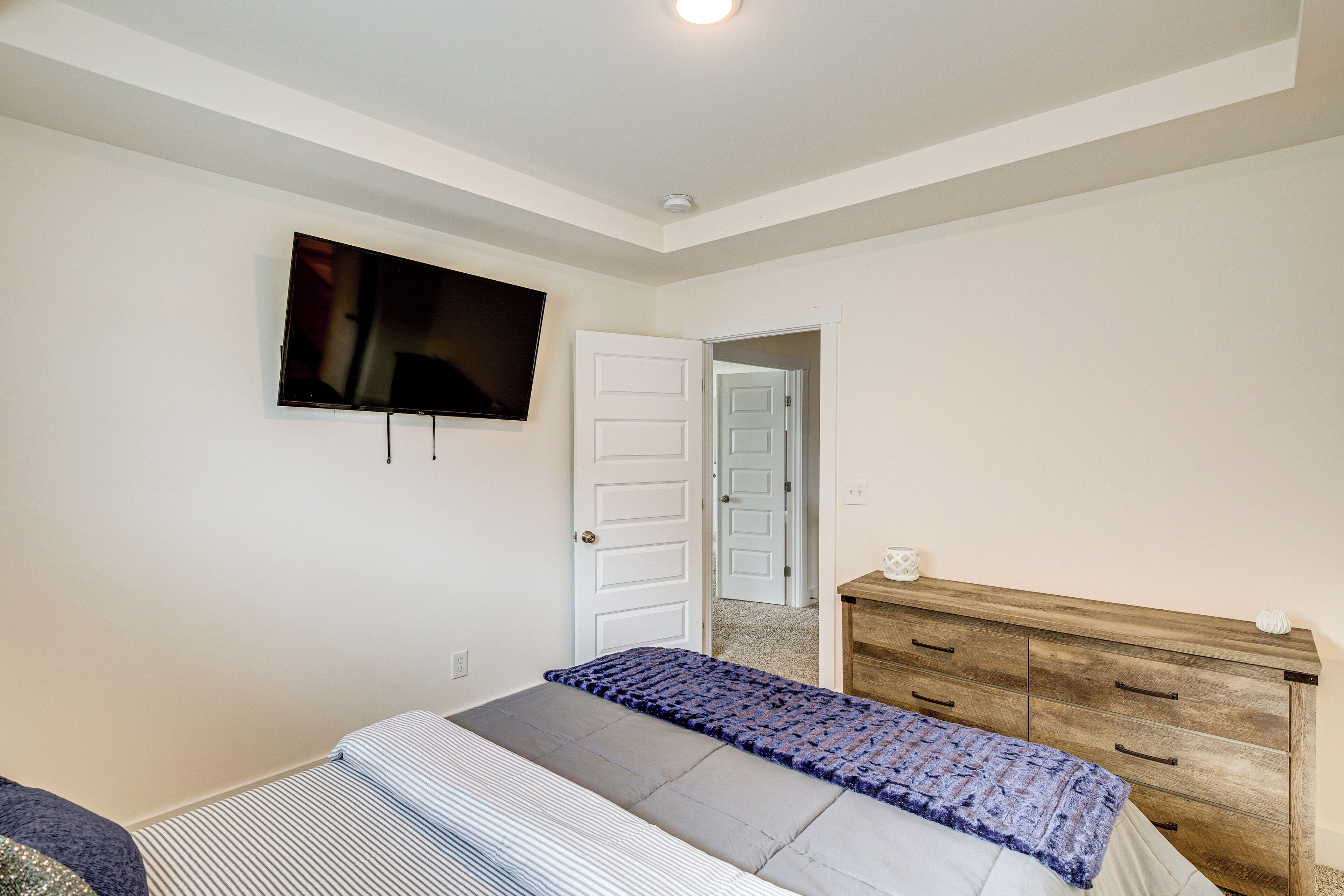 A bedroom with a bed, a wooden dresser, a television on the wall, and a door leading to another room.