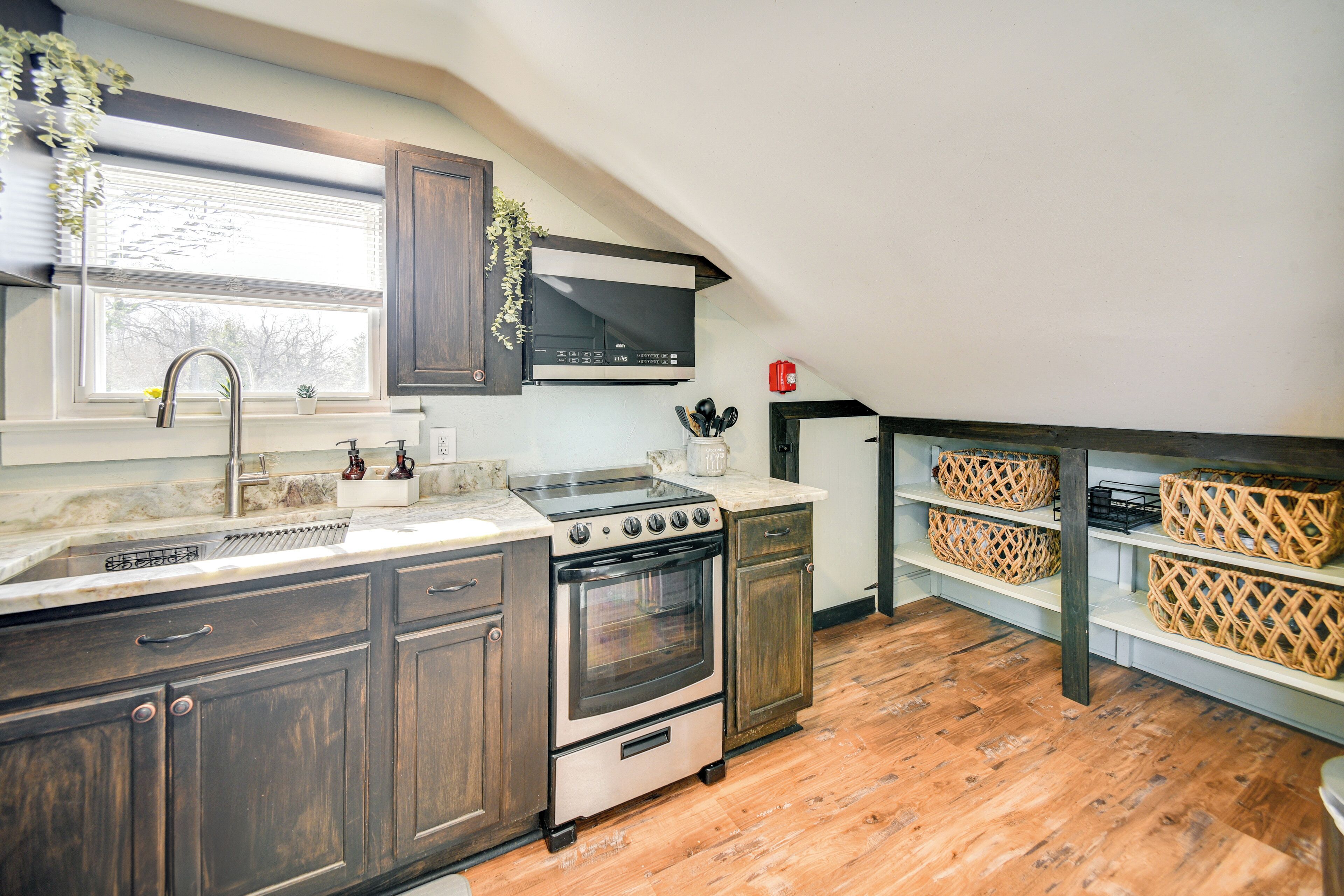 A kitchen with dark wooden cabinets, a stainless steel stove, and a microwave. There is a window with blinds, a sink, and a shelf with baskets.