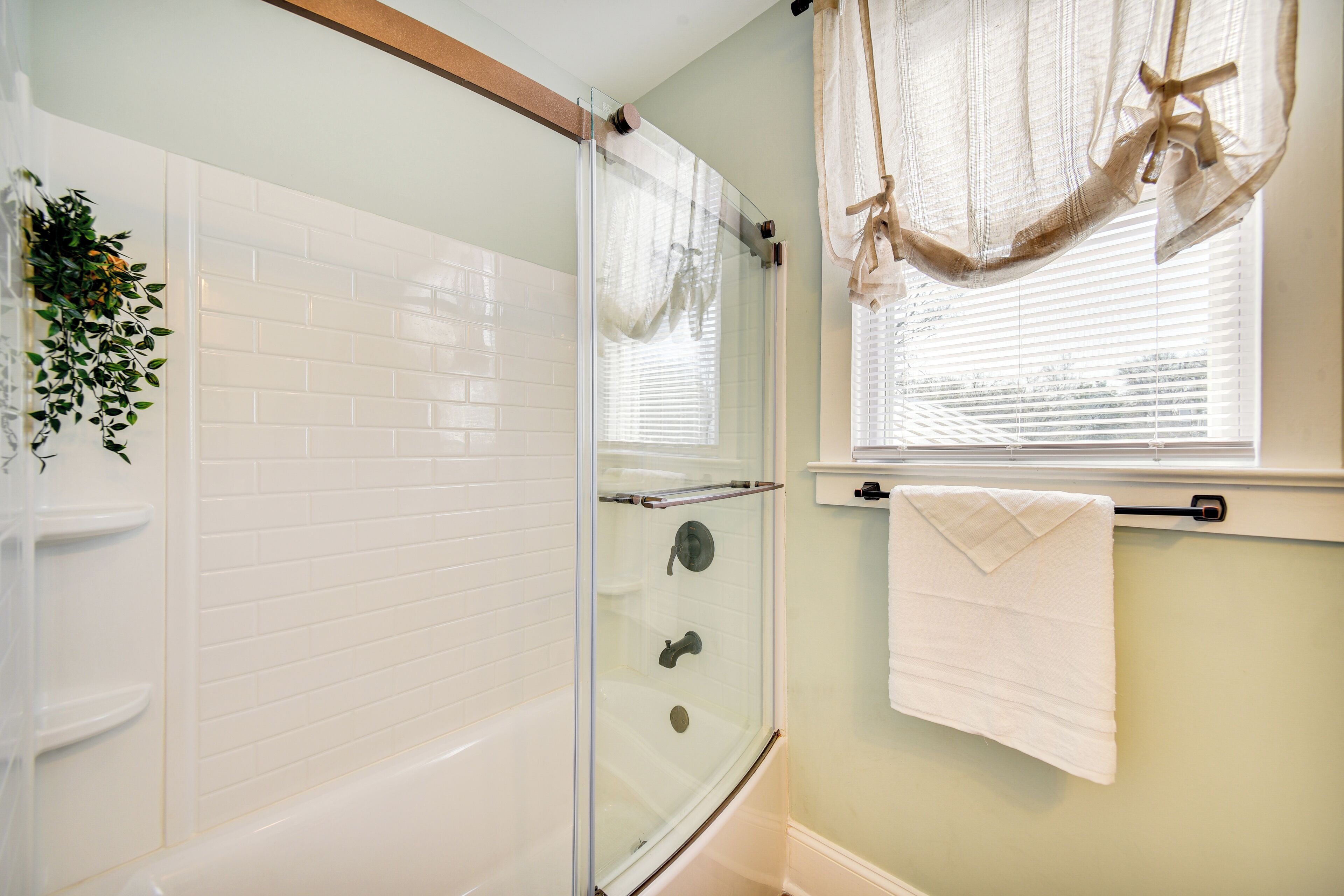 Un baño con ducha de vidrio, paredes con azulejos blancos, una bañera blanca, una ventana con persianas y un perchero con una toalla.