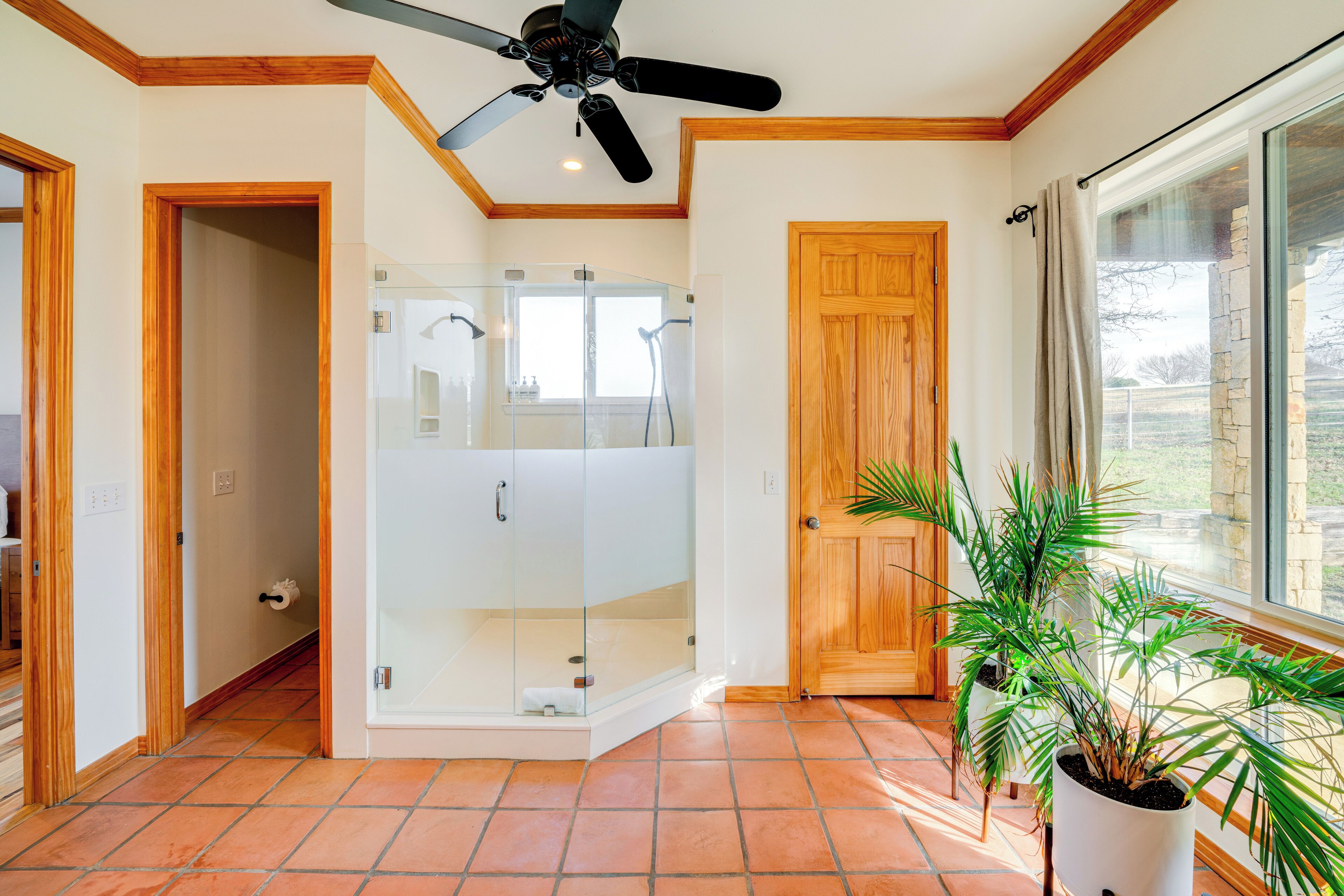 A bathroom with a glass-enclosed shower, wooden doors, a ceiling fan, and a large window.