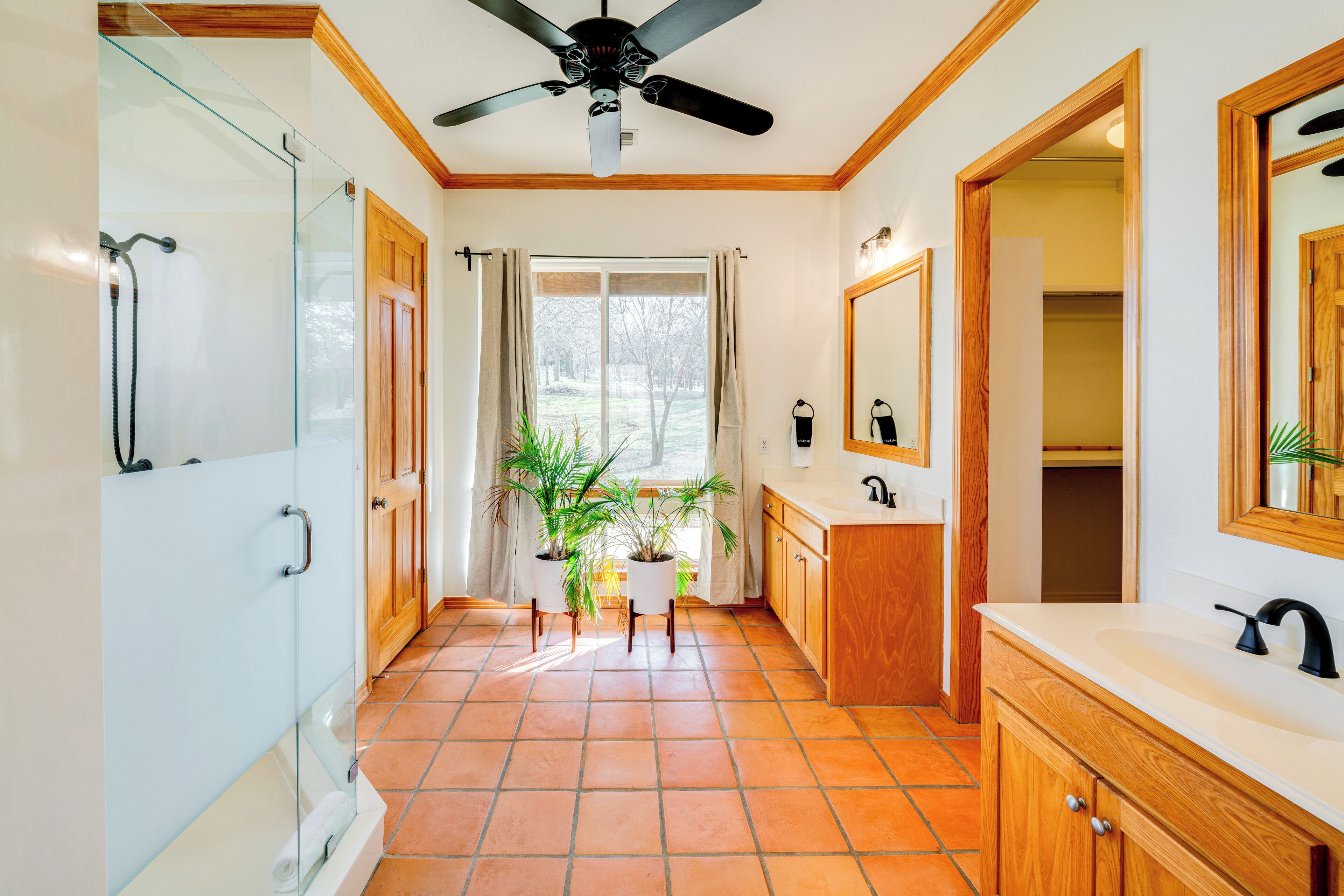 A modern bathroom with a glass shower enclosure, a ceiling fan, a large window with curtains, a sink with a mirror, and wooden cabinetry.