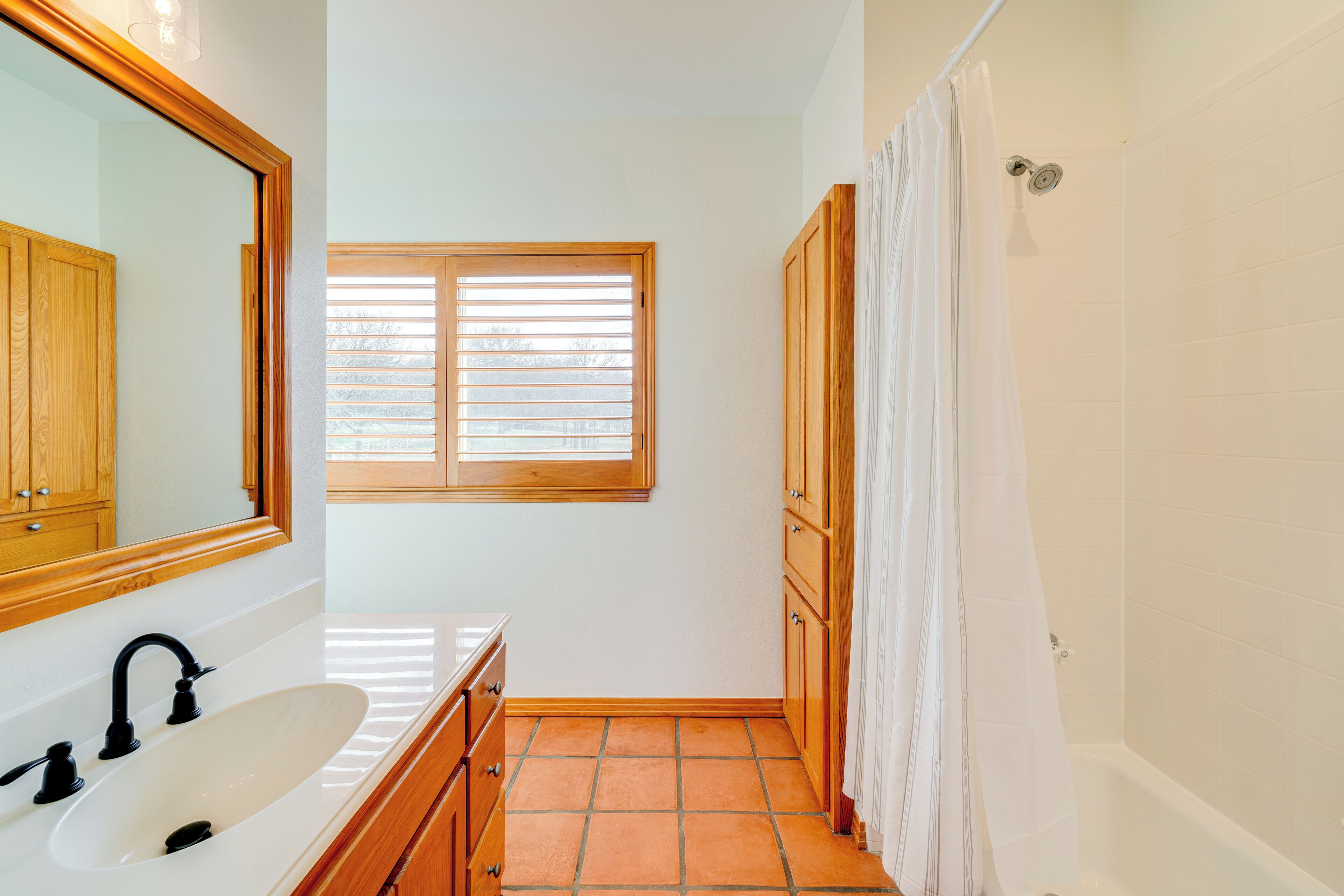 A bathroom with a white sink, wooden cabinets, a window with blinds, and a shower area.