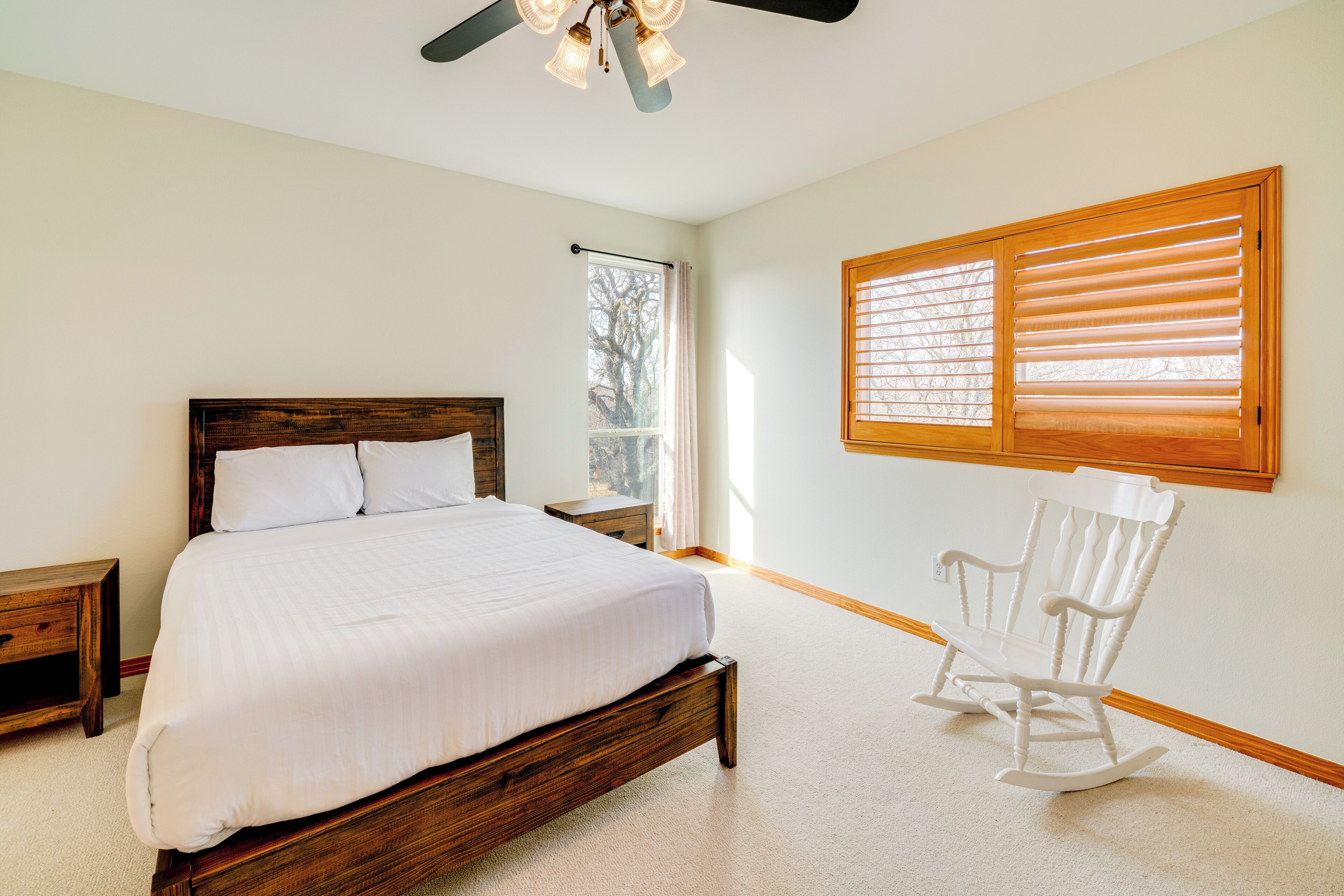 Une chambre à coucher avec un lit en bois, un fauteuil à bascule, une fenêtre avec des stores et un ventilateur de plafond.
