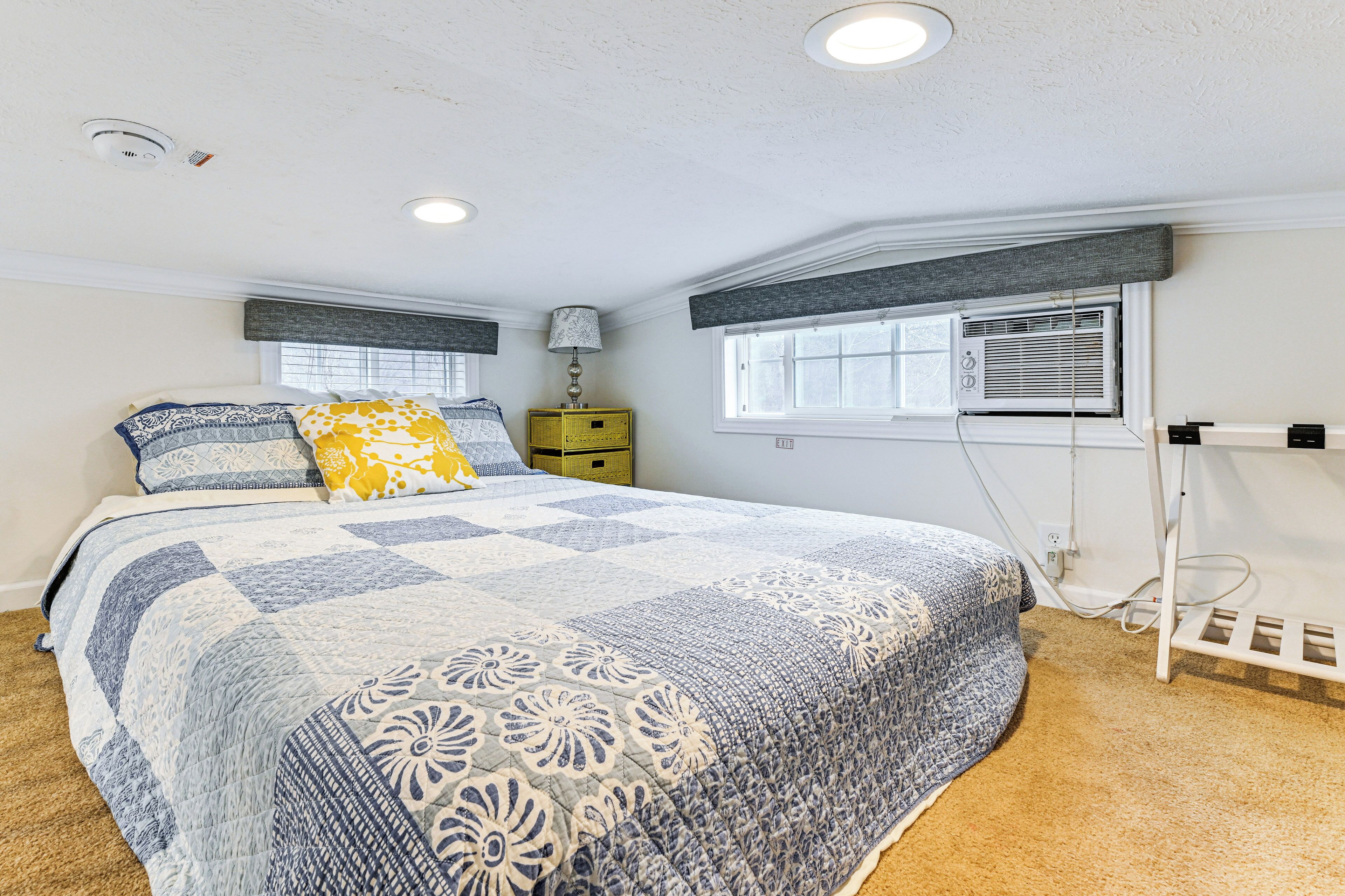 A bedroom with a large bed, a nightstand, a lamp, a window with blinds, and an air conditioning unit.