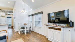 A modern kitchen with white cabinetry, a dining area with a white table and chairs, and a large TV mounted above the counter. - Community Perks + Views: Cozy Lake Norman Cottage (Sherrills Ford)