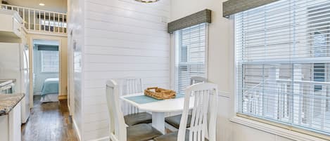 A dining area with a white table and chairs, a chandelier, and large windows with blinds.