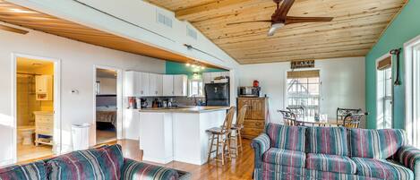 A spacious living area with a wooden ceiling, a sofa set, a dining table, and a kitchen in the background.