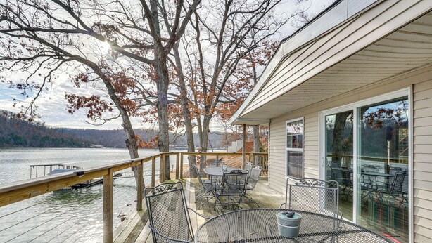 Eine Terrasse mit Gartenmöbeln, die auf einen See blickt, eine Glastür, die zu einer überdachten Terrasse führt, und im Hintergrund Bäume und Berge.
