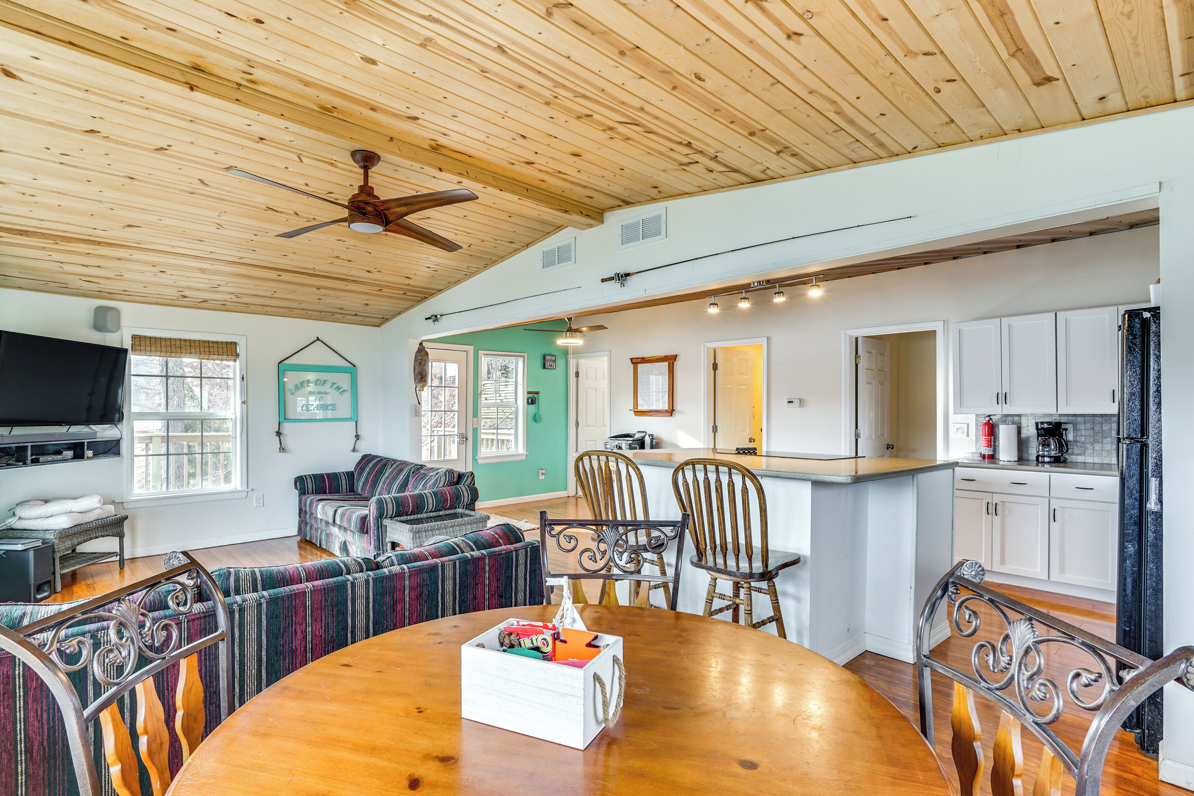 A spacious living room with a wooden ceiling, a dining area with a wooden table and chairs, a kitchen with white cabinets, and a television in the living room.