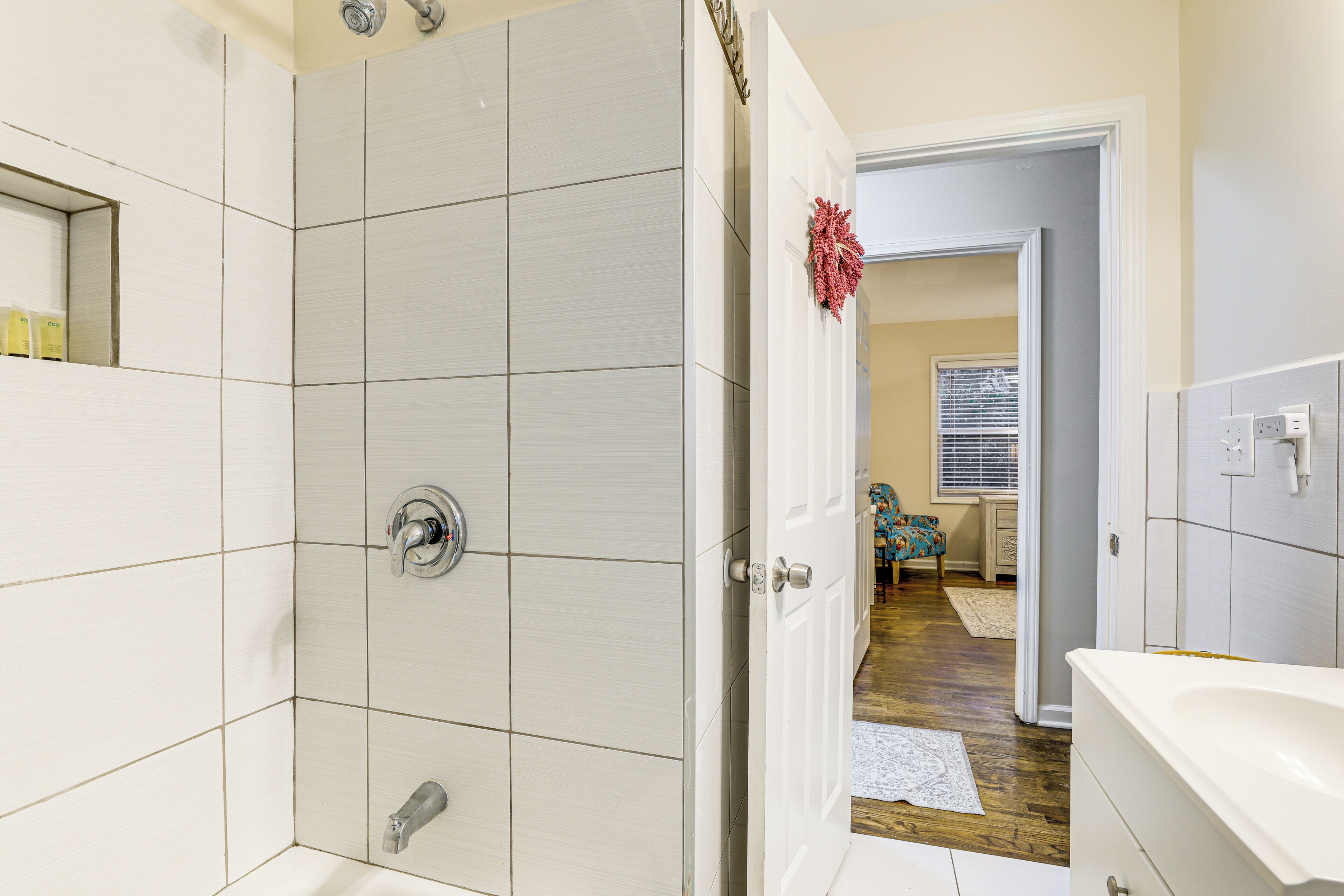 A bathroom with white tiles, a shower, and a door leading to a living area with a blue chair and a window.
