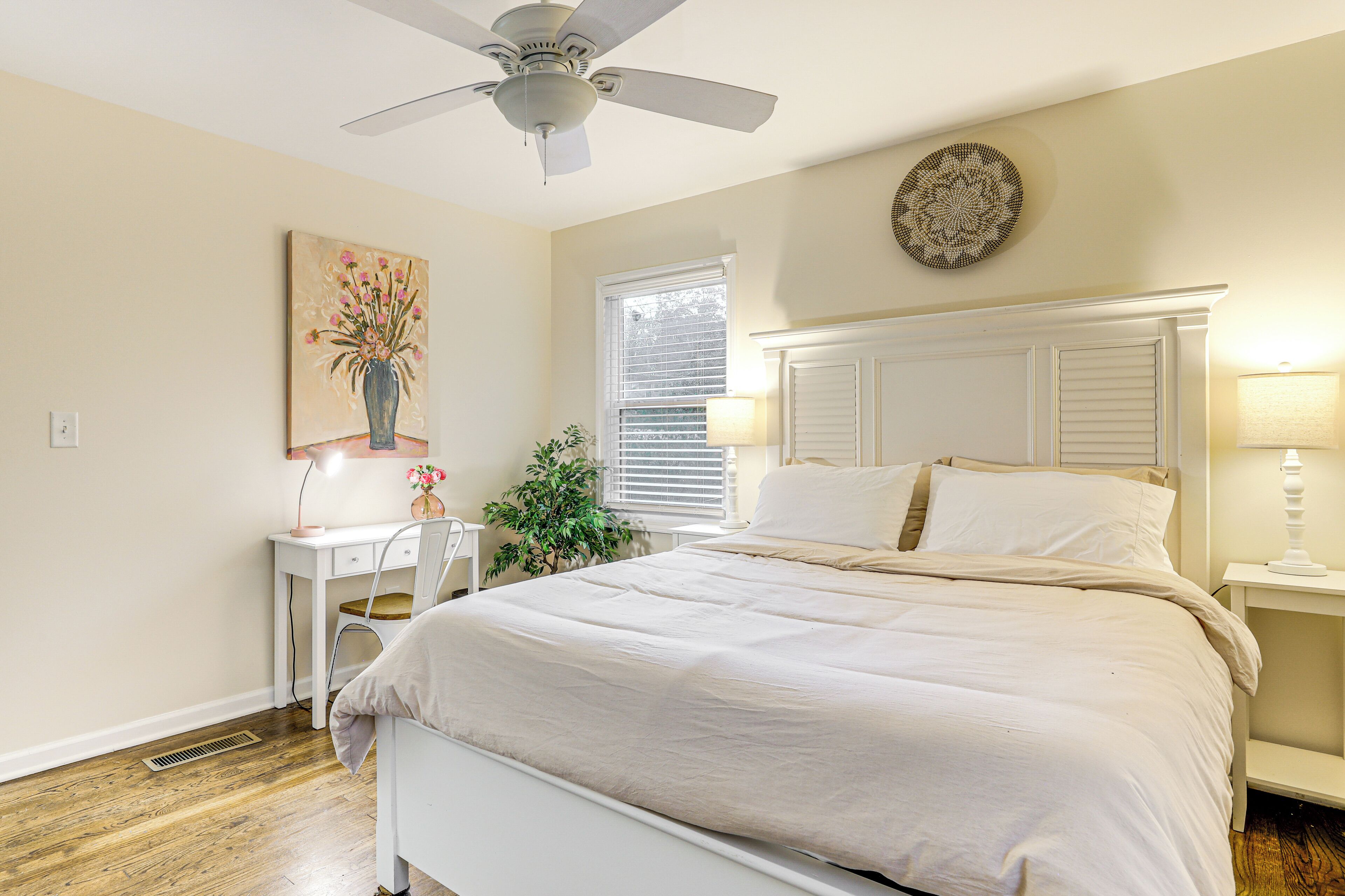 Une chambre à coucher comprenant un lit, un bureau, une chaise, un ventilateur de plafond, une fenêtre avec des stores et un objet décoratif fixé au mur.