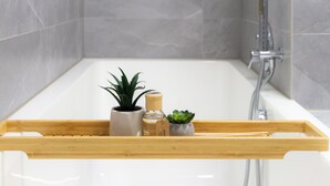 Een moderne badkamer met een bad, grijze tegels, een houten plank met planten en een lijst met een afbeelding aan de muur.