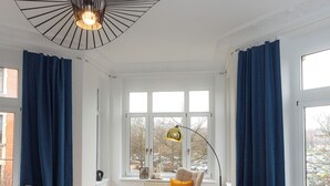 Een moderne woonkamer met een bank, een eettafel met stoelen, een schommelstoel en een groot raam met gordijnen.
