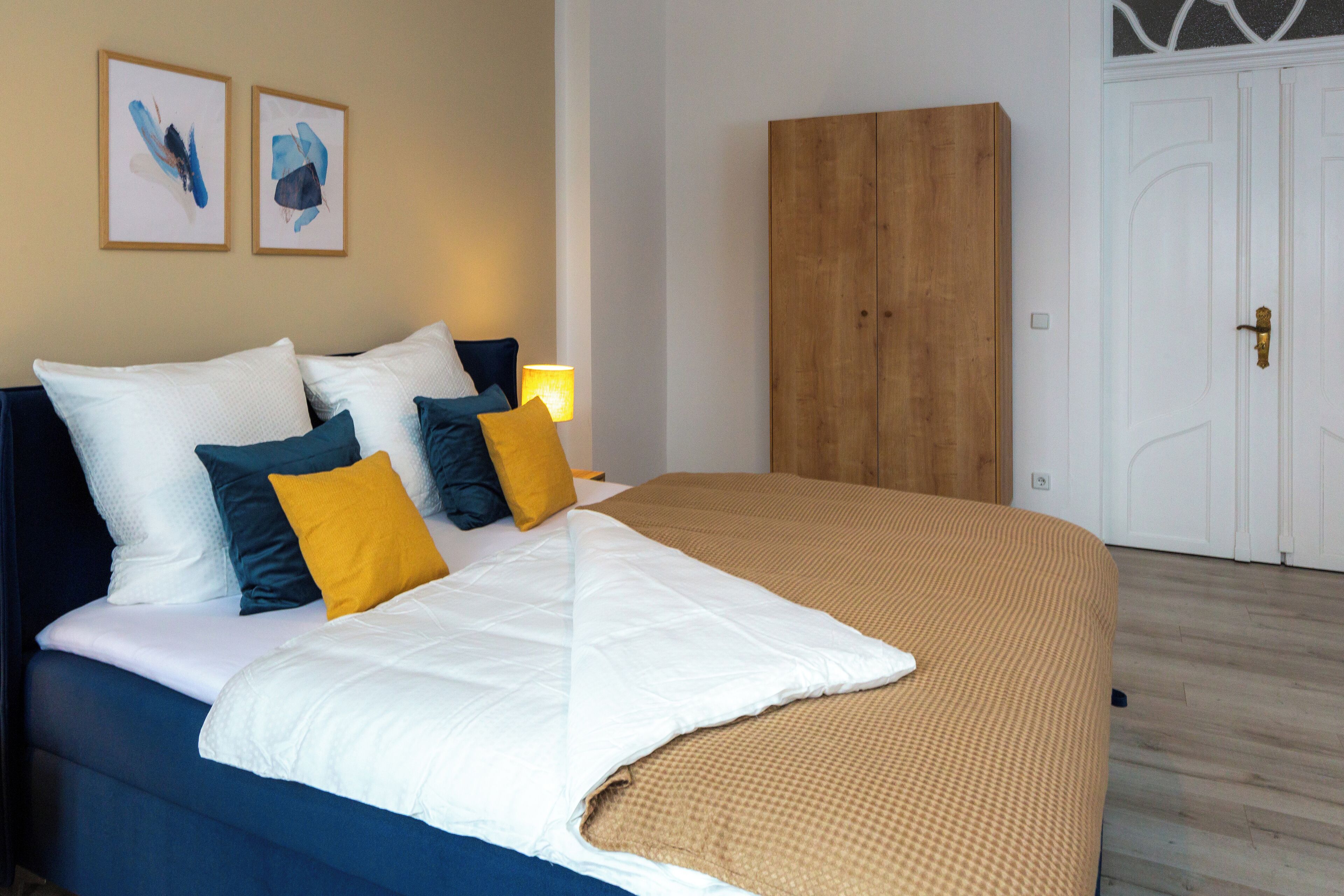 A neatly made bed with white and blue bedding, a wooden wardrobe, and two framed artworks on the wall.