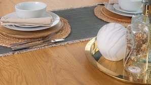 Een eettafel gedekt met borden, kommen en bestek, een vaas met gedroogde planten, en een lamp in een kamer met uitzicht door een raam.