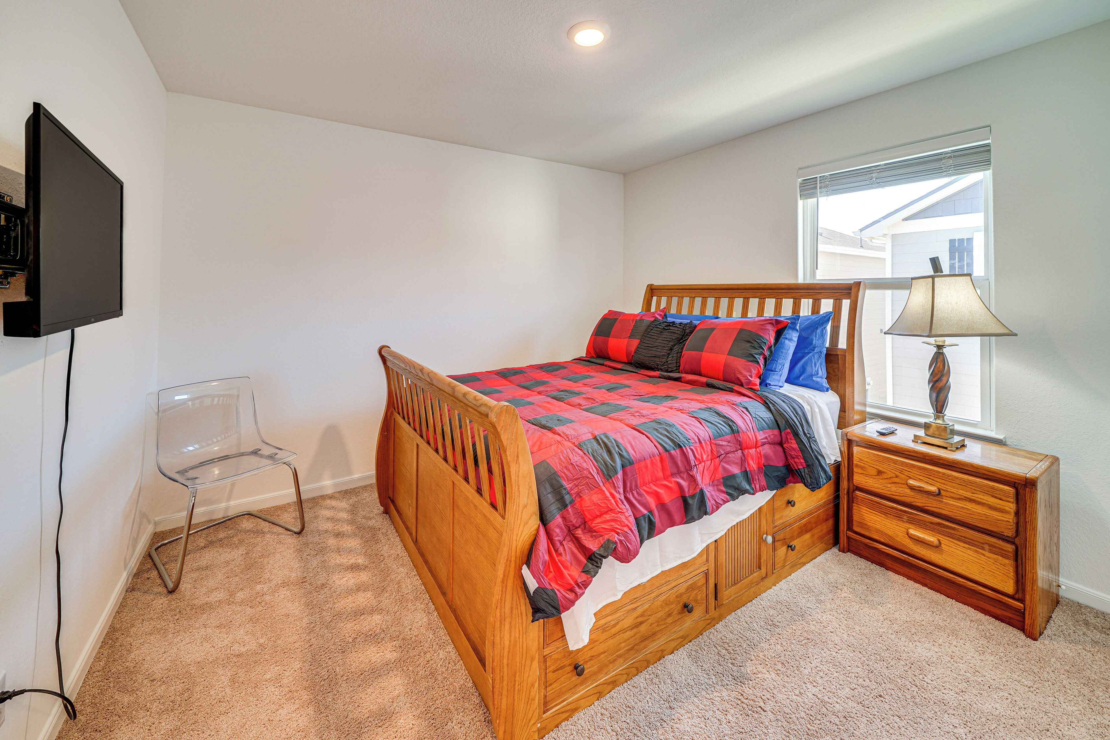 A bedroom with a wooden bed, a nightstand with a lamp, a TV mounted on the wall, and a clear plastic chair.