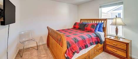 A bedroom with a wooden bed, a nightstand with a lamp, a TV mounted on the wall, and a clear plastic chair.