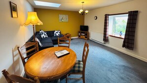 Una sala de estar acogedora con un sofá, una mesa de comedor y sillas. Hay un televisor, un reloj y una ventana con cortinas.