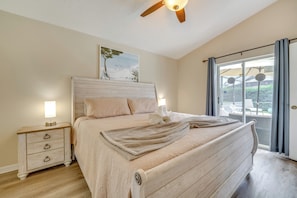 A bedroom with a bed, a nightstand, a ceiling fan, a window with curtains, and a view of the outdoors.