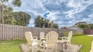 Un jardin avec une clôture en bois, une pelouse et un espace patio aménagé avec des chaises et une table en plastique blanc.