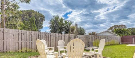 A backyard with a wooden fence, a lawn, and a patio area with white plastic chairs and a table.