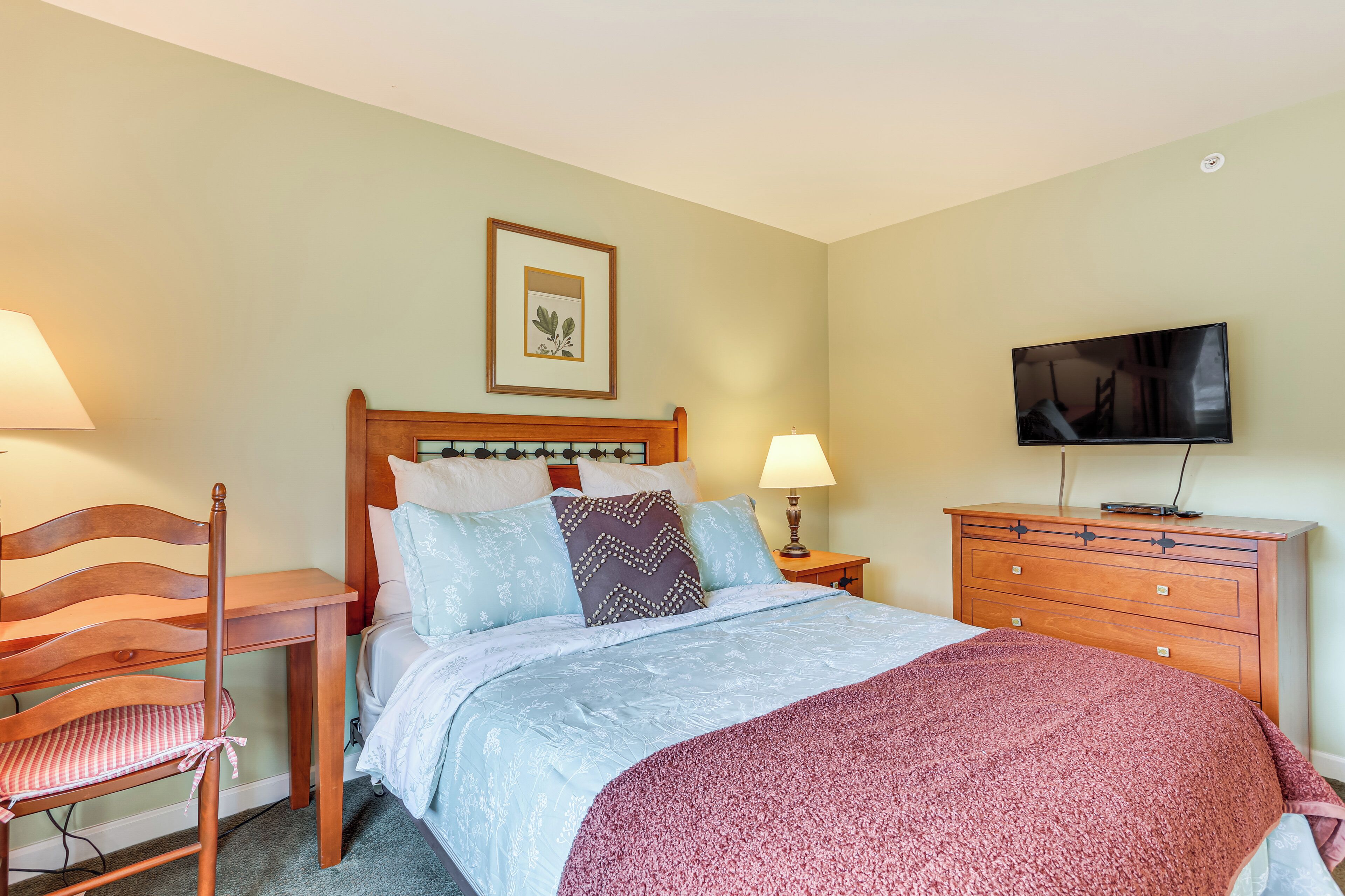 A neatly arranged bedroom with a bed, a wooden chair, a dresser, a television, and a framed picture on the wall.