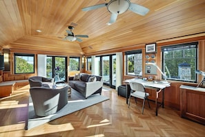 A spacious room with a wooden ceiling, large windows, a ceiling fan, a desk with a lamp, and a comfortable seating area.