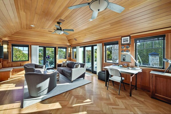 A spacious room with a wooden ceiling, large windows, a ceiling fan, a desk with a lamp, and a comfortable seating area.