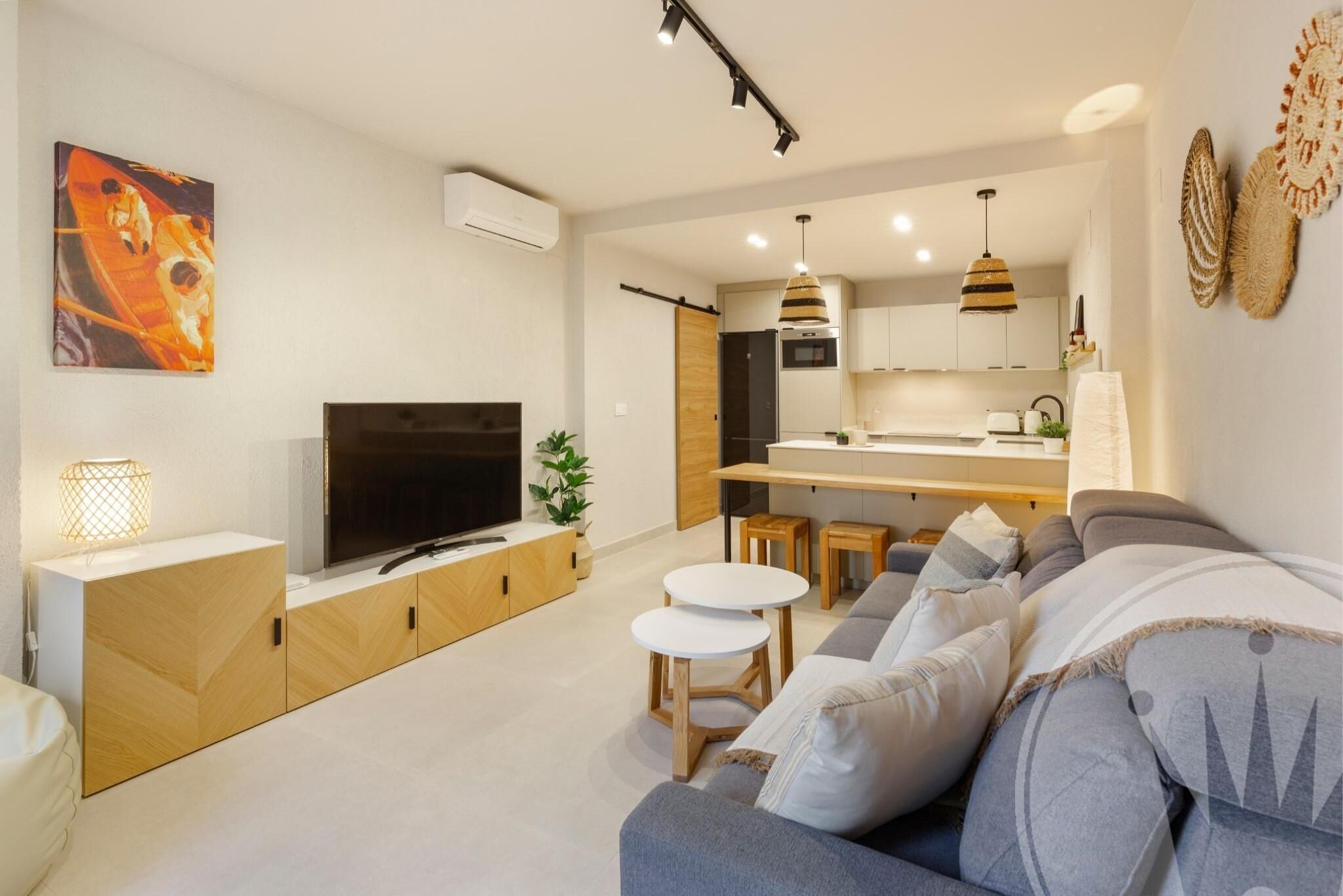 Una sala moderna con sofá, televisión y mesitas. Cuenta con una cocina con refrigerador y una mesa de comedor.