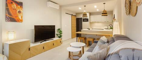 Una sala moderna con sofá, televisión y mesitas. Cuenta con una cocina con refrigerador y una mesa de comedor.