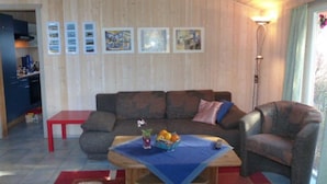 A living room with a wooden ceiling, a sofa, a chair, a coffee table with a blue cloth, and a small table with a plant and fruit. - Ferienhaus mit Sauna im Ferienpark Mirow (Mirow)