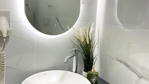 Un baño con un lavamanos blanco, un espejo redondo, un secador de pelo de mano y una planta en maceta.
