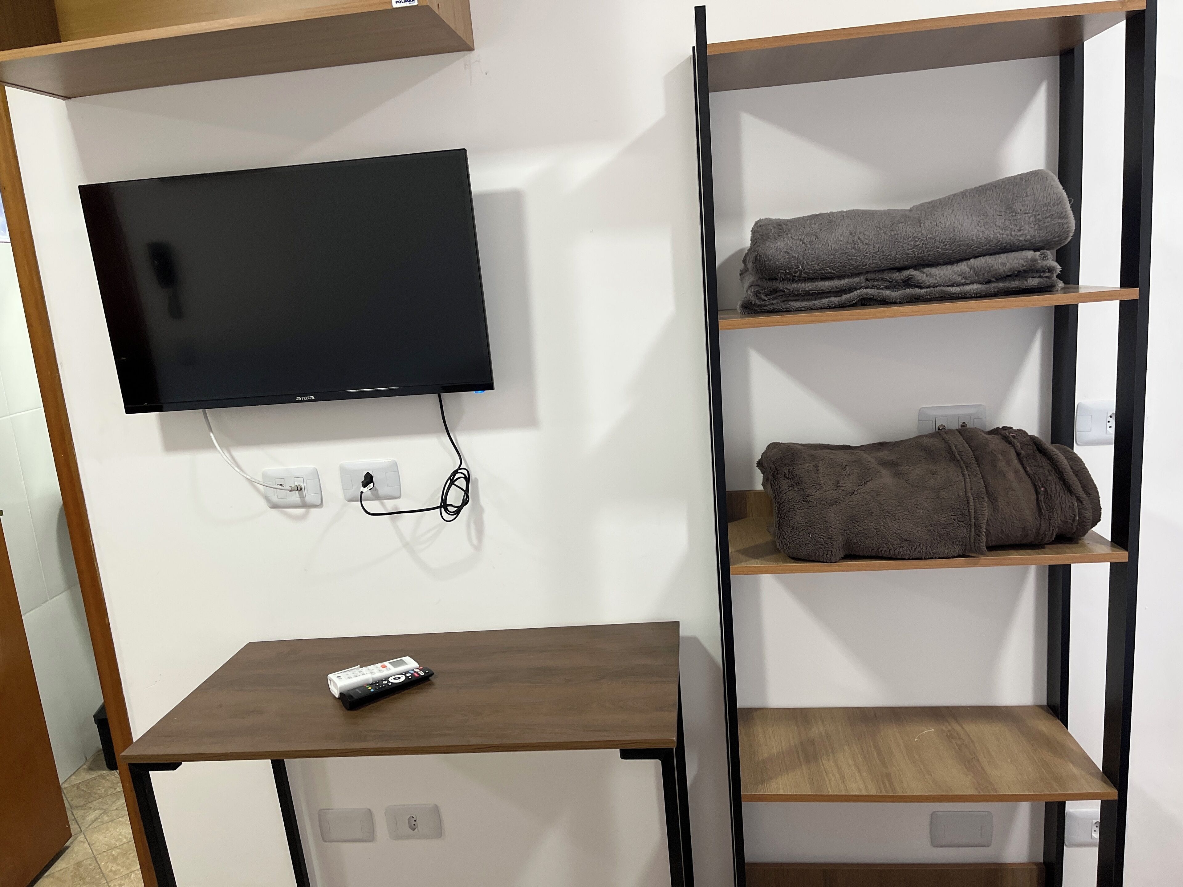 A modern room with a flat-screen TV mounted on the wall, a wooden table with a remote control, and a built-in shelving unit with folded towels.