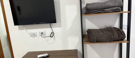 A modern room with a flat-screen TV mounted on the wall, a wooden table with a remote control, and a built-in shelving unit with folded towels.