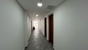 A long, narrow hallway with white walls and grey tiled flooring. There are doors on both sides, and a fire extinguisher is visible on the left wall. - CONDOMÍNIO RESIDENCIAL VITÓRIA 3 (São Paulo)