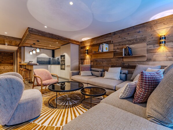 Un salon moderne avec un canapé, des fauteuils et une table basse. On y trouve également un coin cuisine avec un réfrigérateur et un mur en bois doté d’étagères.