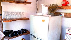 Een kleine keuken met een witte koelkast, planken met glazen en zwarte bekers, en een rode waterkoker op een houten plank.